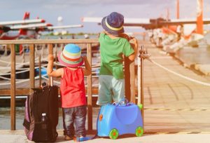 Two children on vacation looking at sea planes while carrying their luggage.